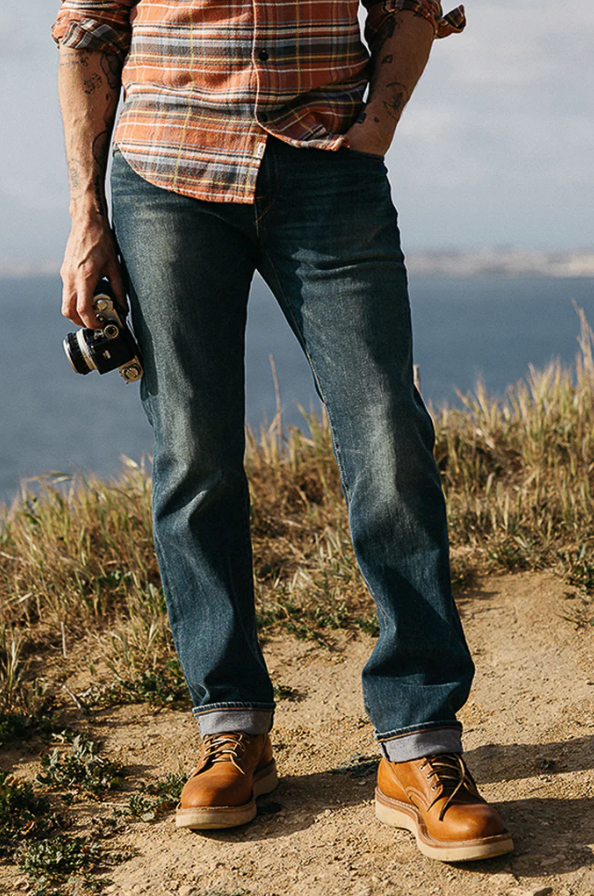Person wearing blue jeans and brown shoes standing on a dirt path with a scenic background.