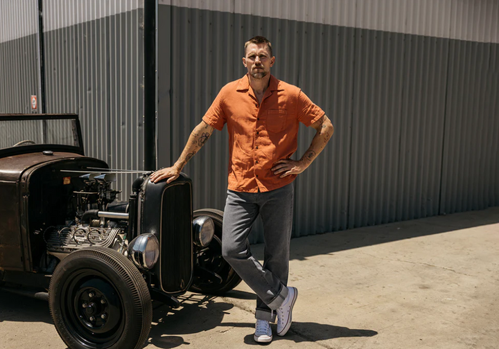 Man in an orange shirt standing next to a vintage car with a metallic wall in the background