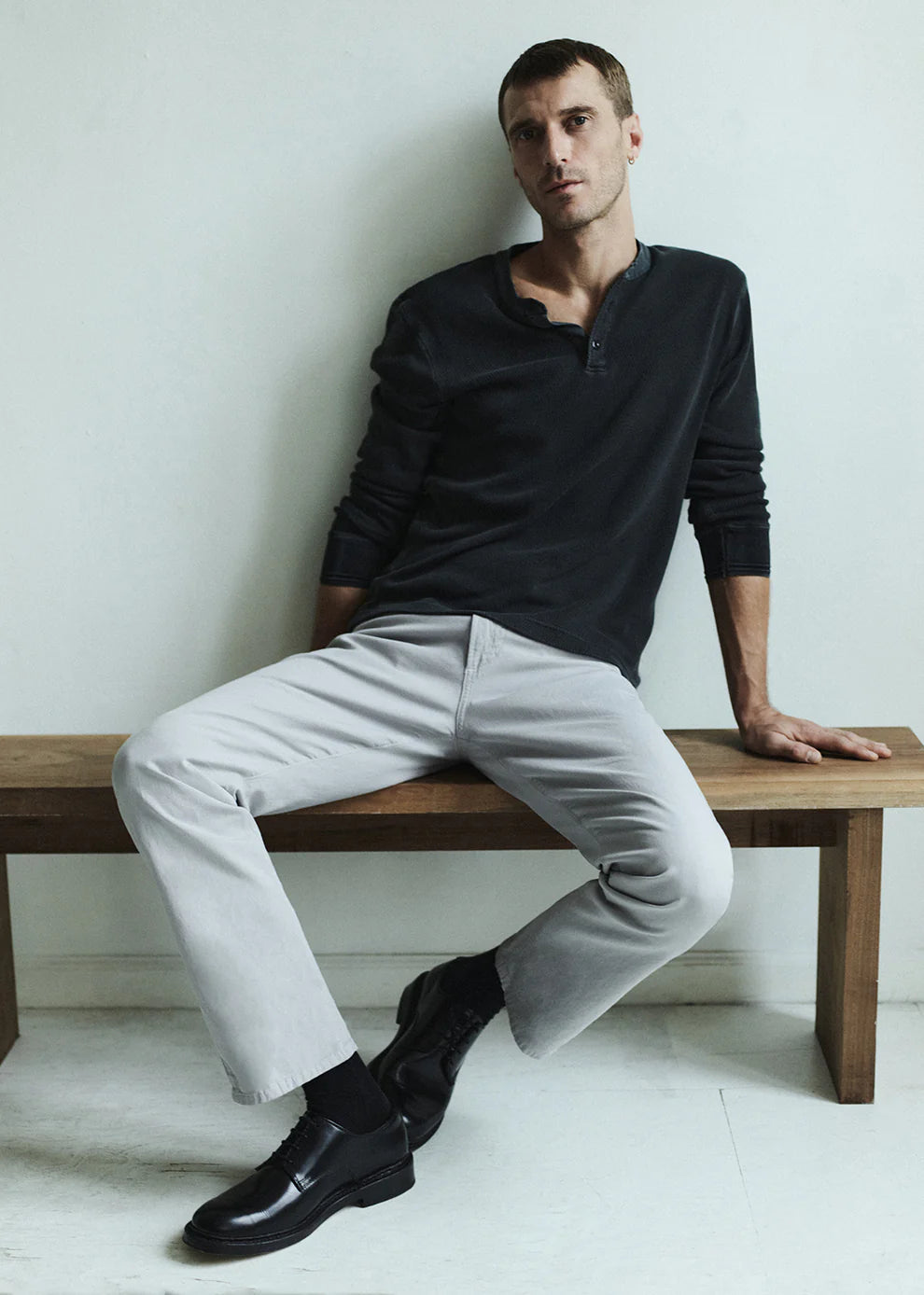 Man sitting on a wooden bench wearing a black shirt, light gray pants, and black shoes.