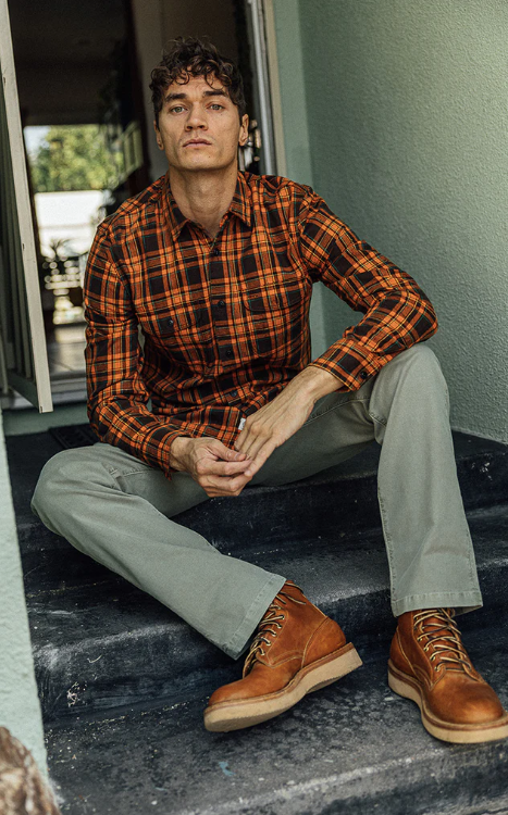 Man sitting on steps wearing a plaid shirt, beige pants, and brown boots.