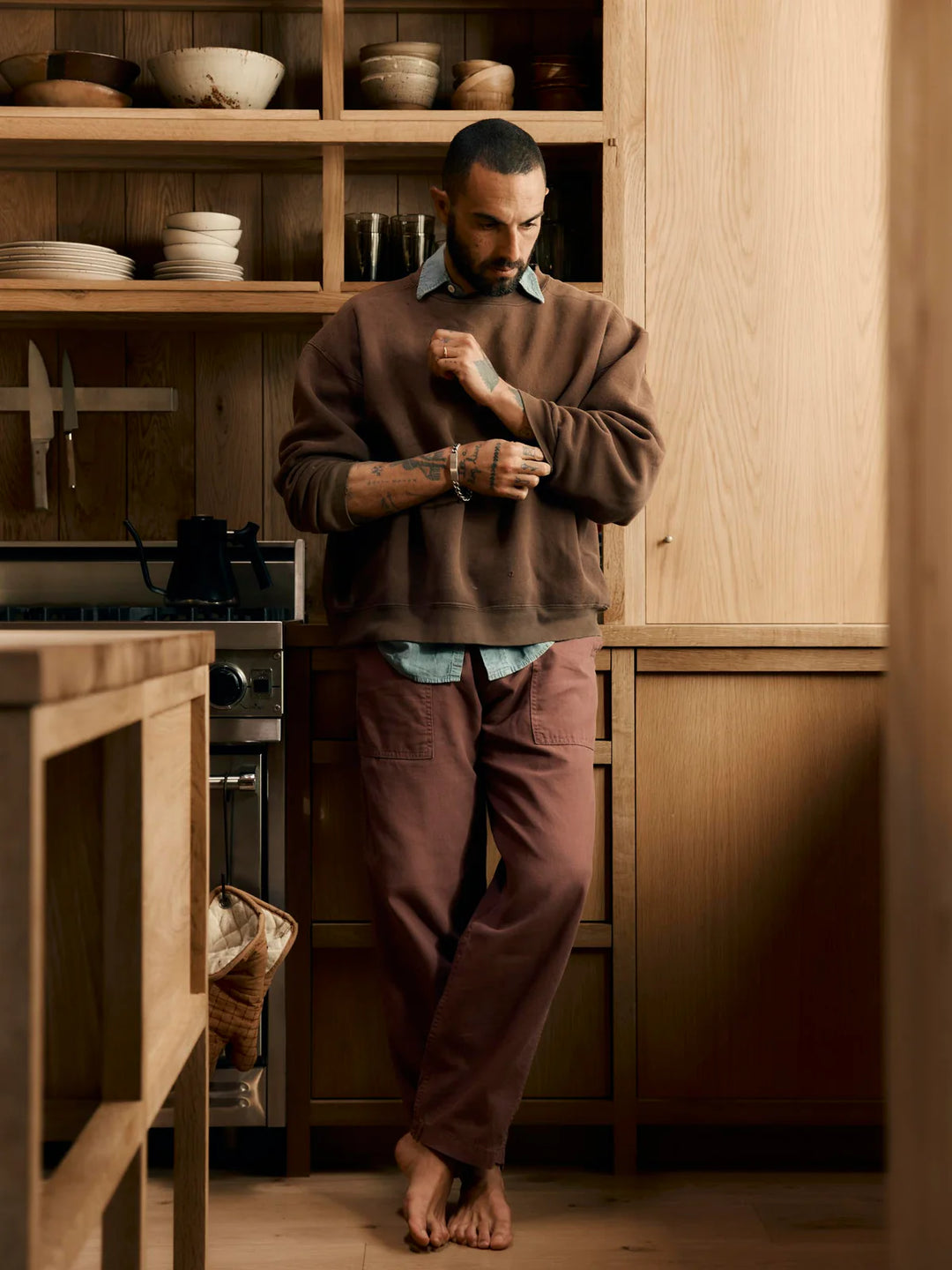 Man standing in a kitchen wearing a brown sweater and pants.