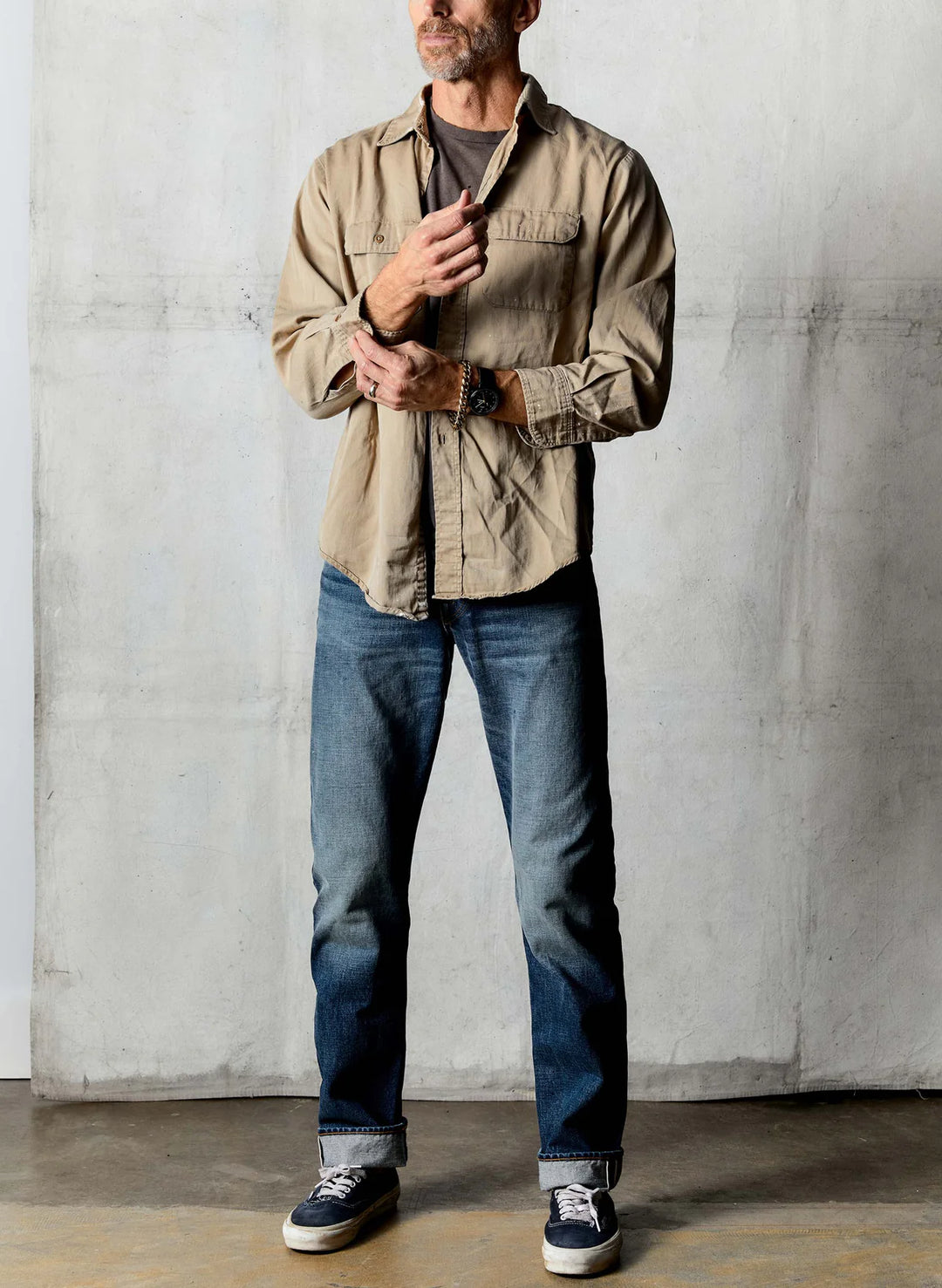 Man wearing a beige jacket, dark shirt, blue jeans, and black shoes against a plain wall.