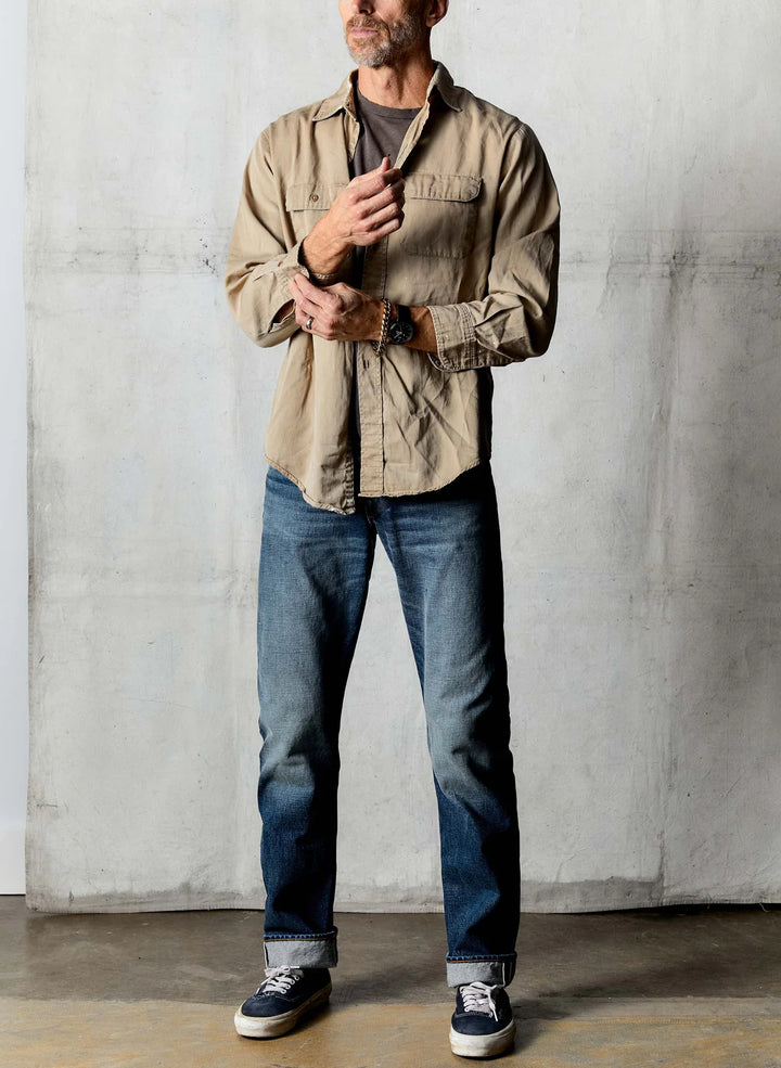 Man wearing a beige jacket, dark shirt, blue jeans, and black shoes against a plain wall.