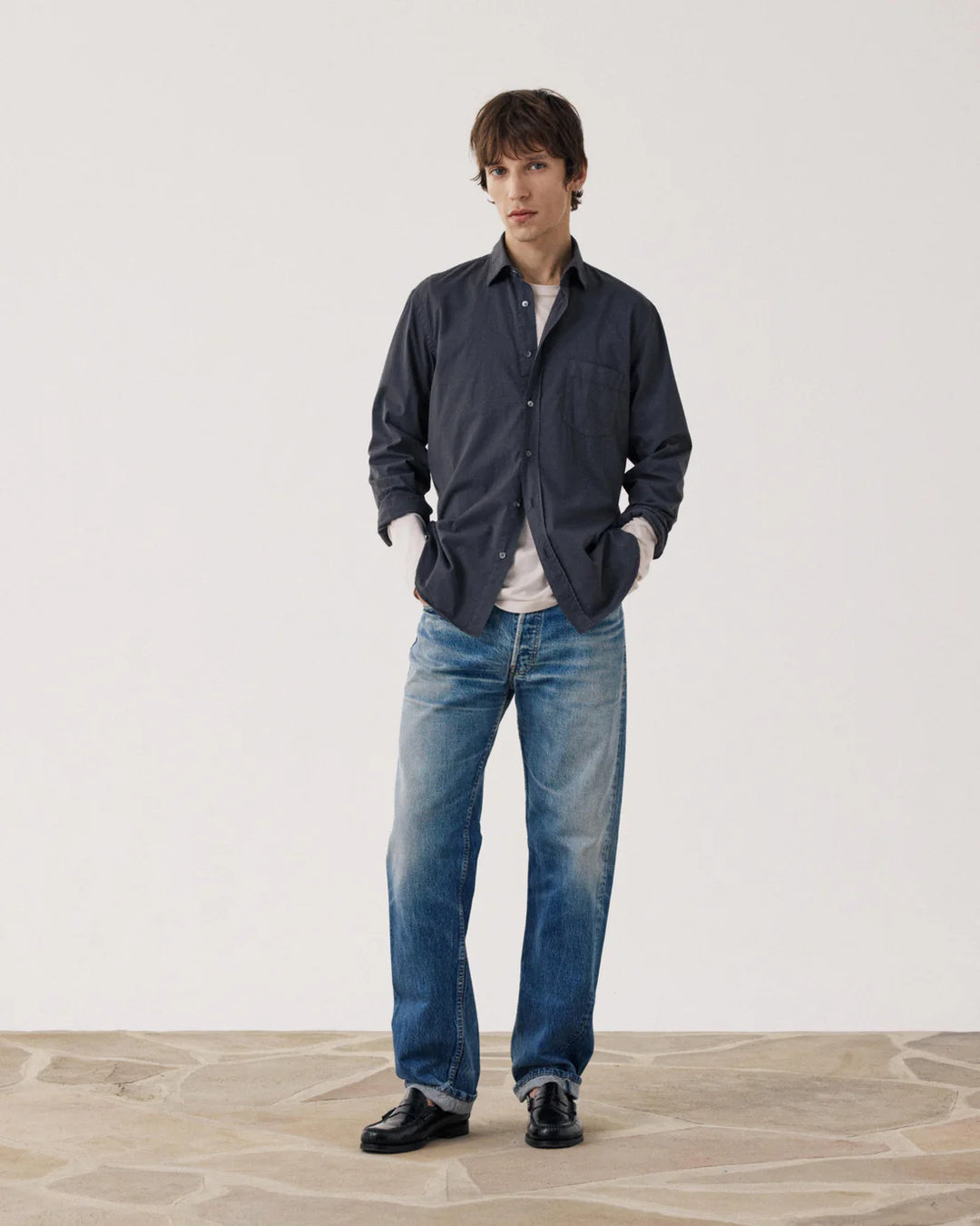 Man wearing a dark jacket, light shirt, and blue jeans standing against a plain background