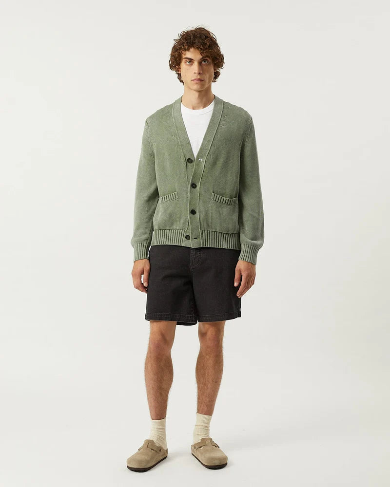 Man wearing a green cardigan over a white shirt with black shorts on a white background