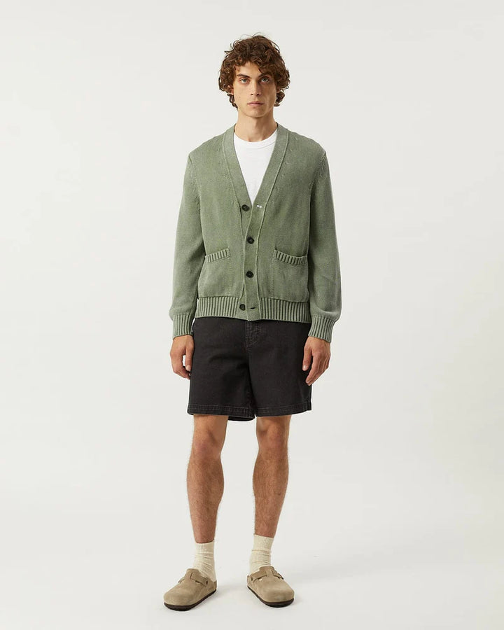 Man wearing a green cardigan over a white shirt with black shorts on a white background
