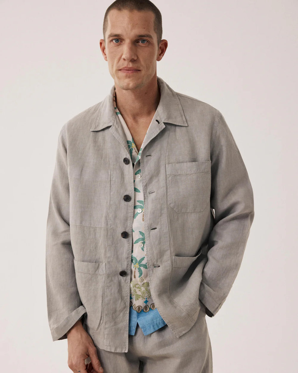 Man wearing a light gray jacket over a patterned shirt on a plain background