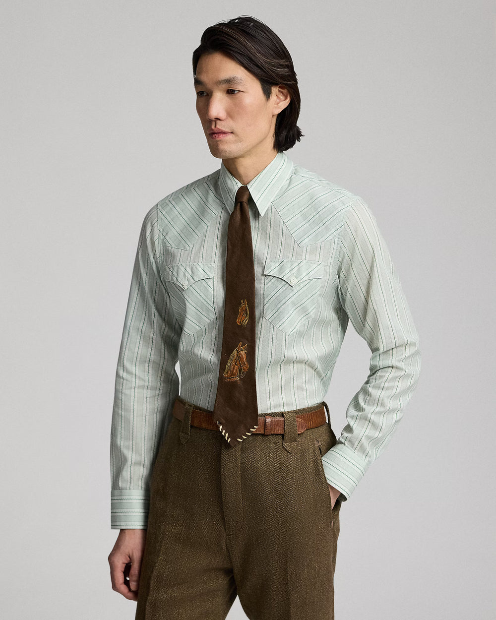 Man wearing a light green striped shirt with a brown tie and pants on a gray background