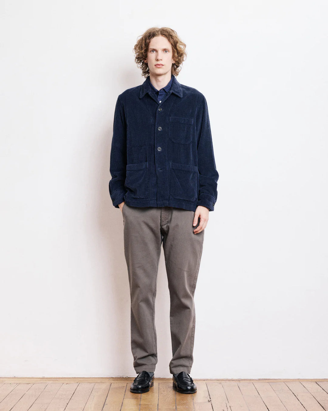 Man wearing a navy jacket and gray pants standing against a white wall.