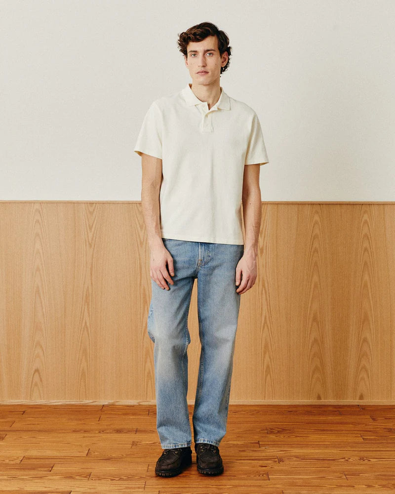 Man wearing a white polo shirt and blue jeans standing in a room with wooden floor and wall.