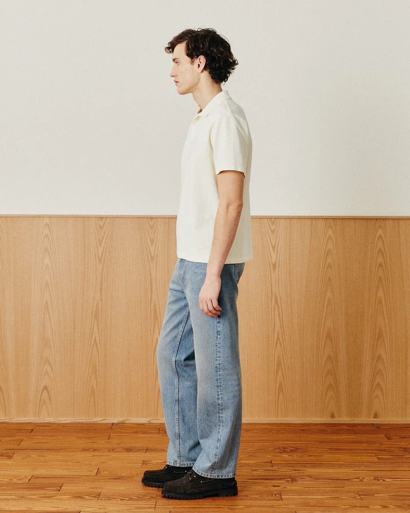 Man wearing a white shirt and blue jeans standing in a room with wooden flooring and paneling.