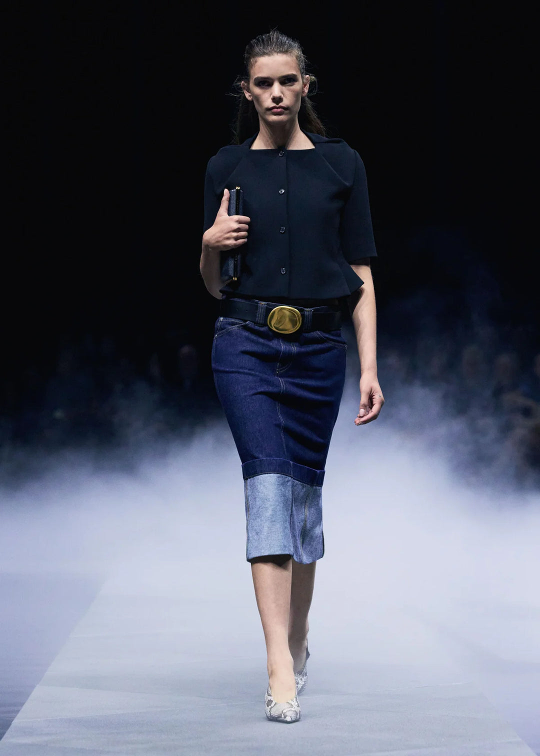 Model walking on a runway wearing a black top and blue denim skirt with a belt.