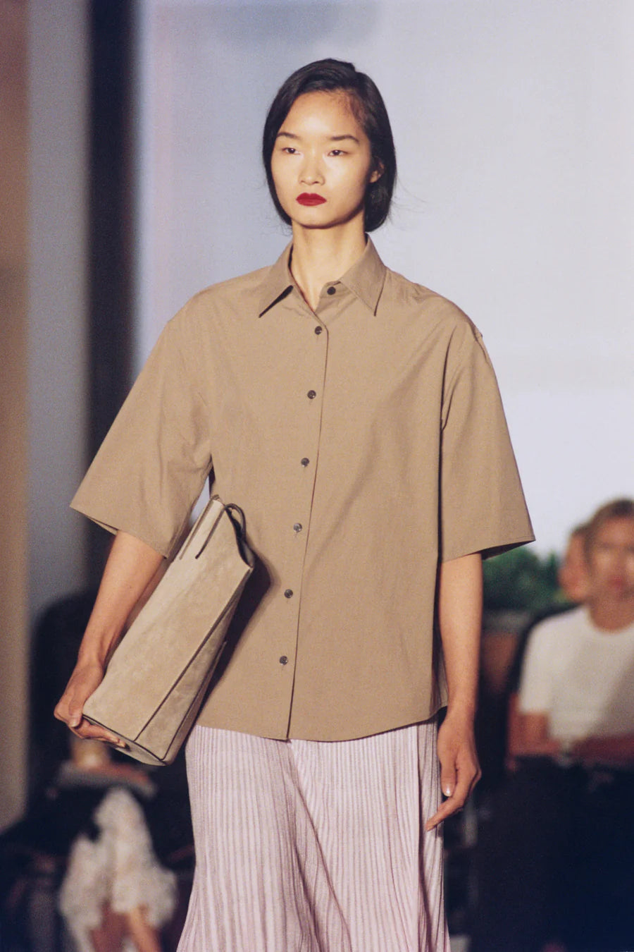 Model wearing a beige shirt and striped pants holding a clutch on a runway.