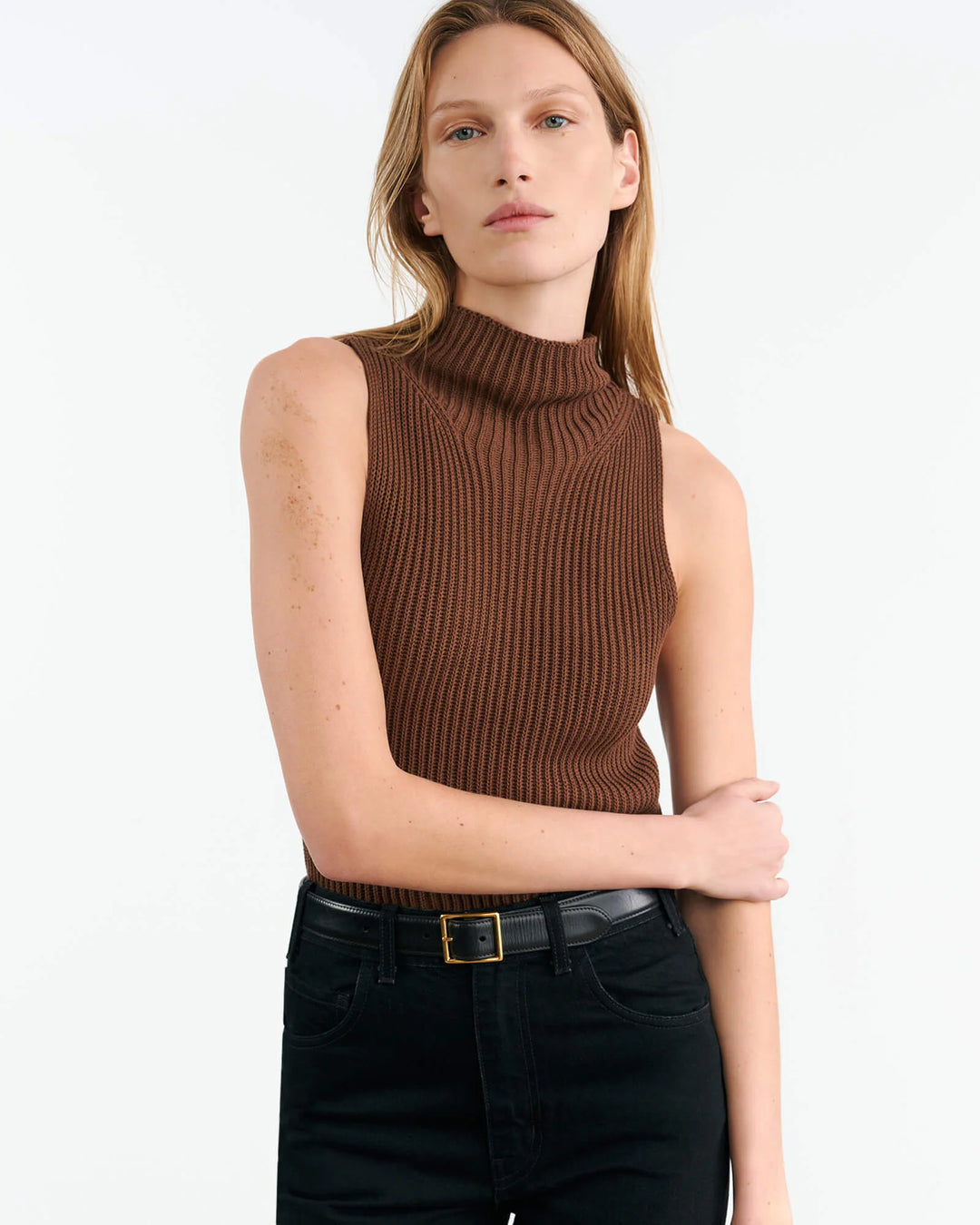 A woman wearing a brown sleeveless mock neck sweater by Nili Lotan. She is also dressed in black trousers and there is a visible white shirt underneath the sweater.