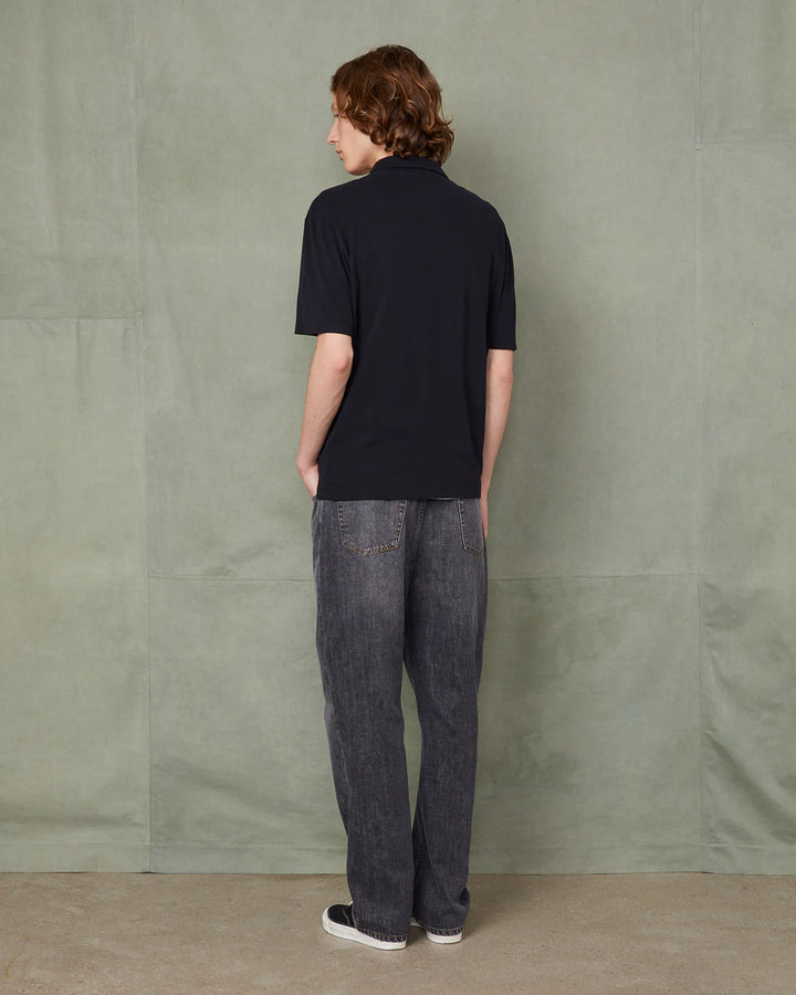 Person wearing a black polo shirt and dark jeans standing against a textured green wall.