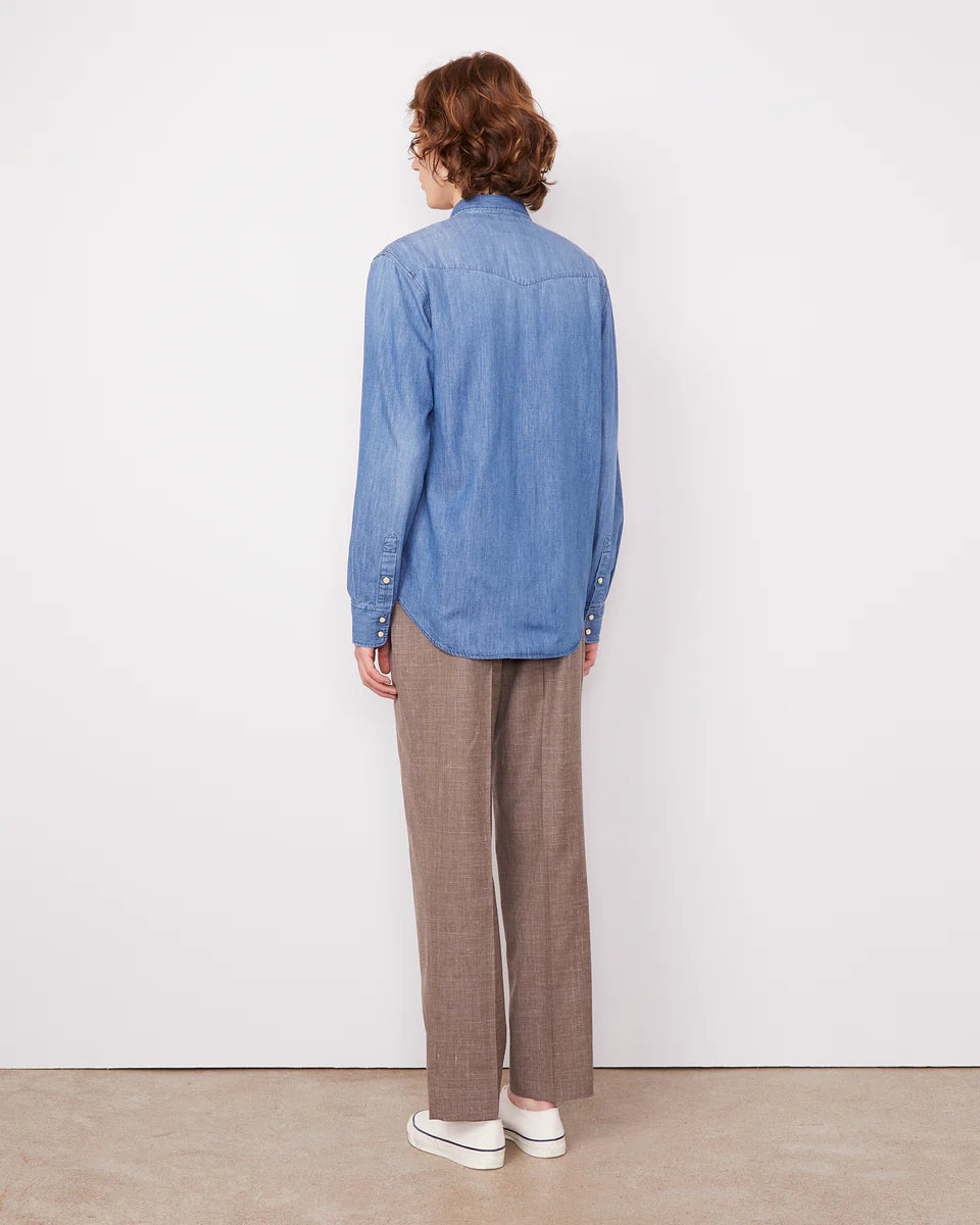 Person wearing a blue denim shirt and brown plaid pants standing against a white wall.