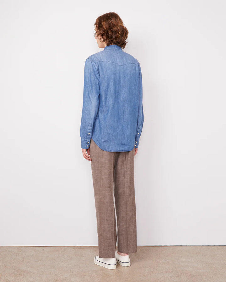 Person wearing a blue denim shirt and brown plaid pants standing against a white wall.