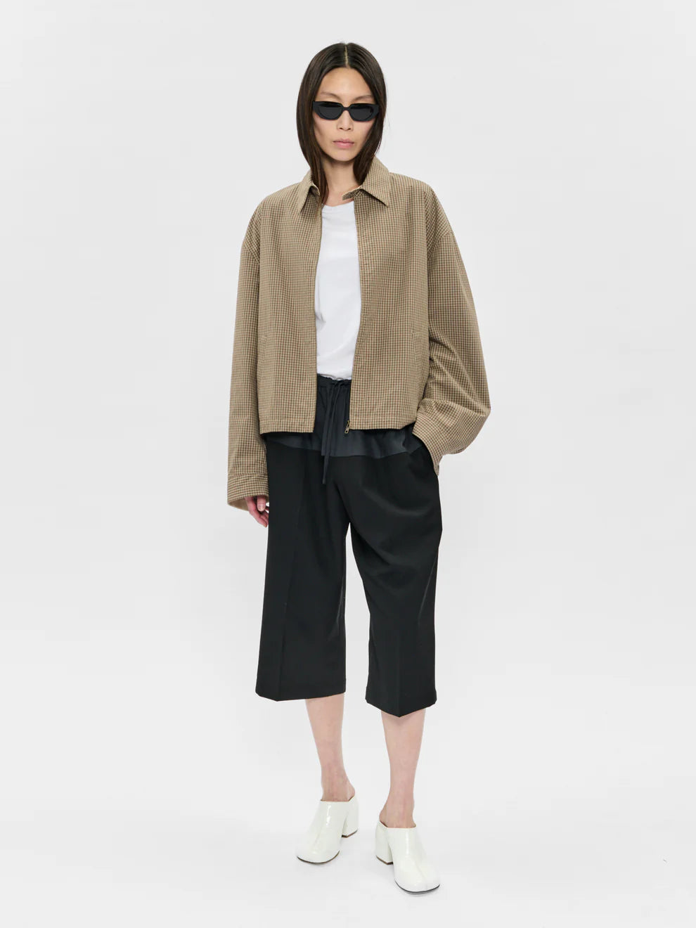 Person wearing a beige jacket, white shirt, black pants, and sunglasses on a white background