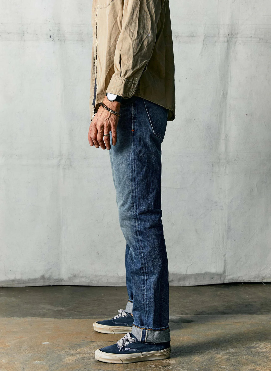 Person wearing a beige shirt, dark jeans, and blue sneakers against a gray concrete wall.