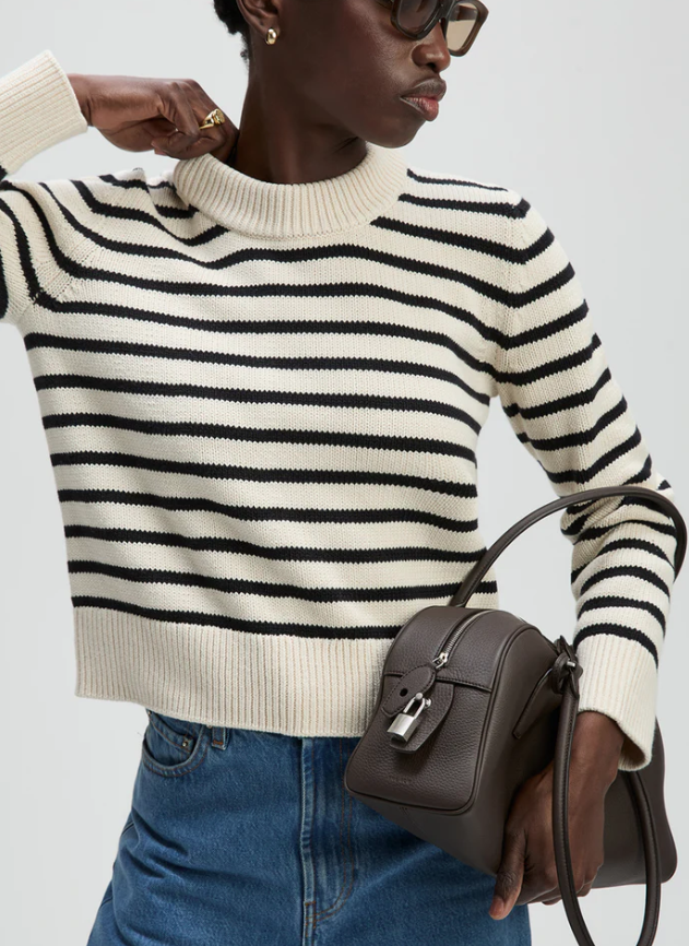 Person wearing a black and white striped sweater holding a brown handbag against a light gray background