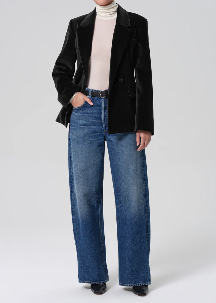 Person wearing a black blazer, beige turtleneck, and blue jeans on a white background