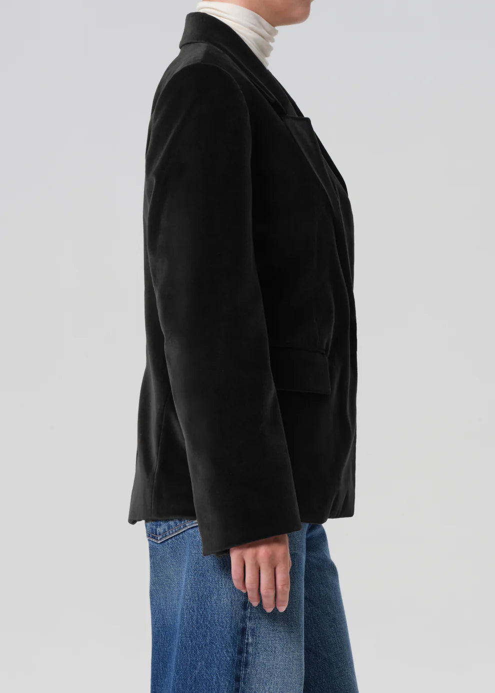 Person wearing a black coat over a white turtleneck and blue jeans on a light gray background