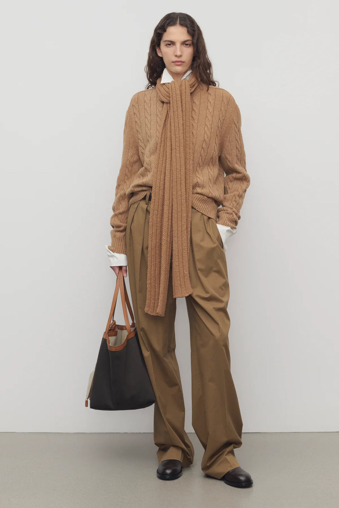 Person wearing a brown outfit with a scarf and holding a black bag against a white background