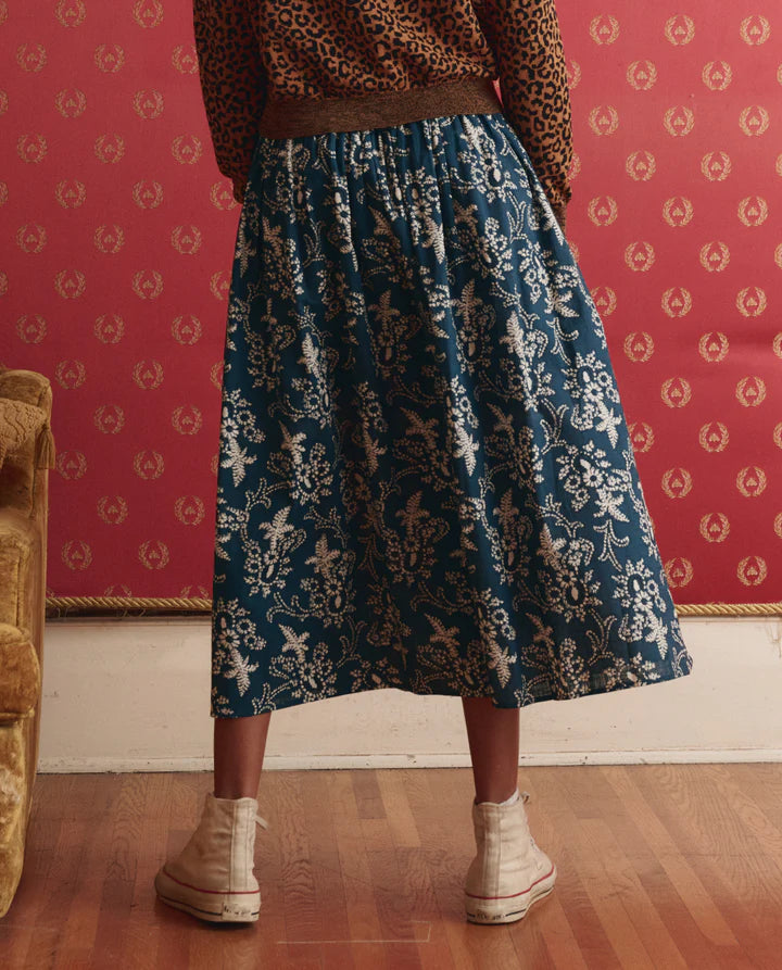 Person wearing a brown sweater and blue floral skirt standing against a patterned wall.