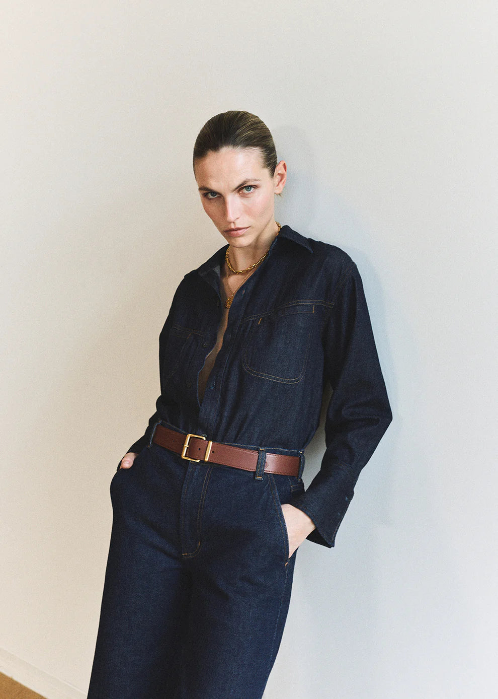 Person wearing a dark denim outfit with a brown belt against a plain background