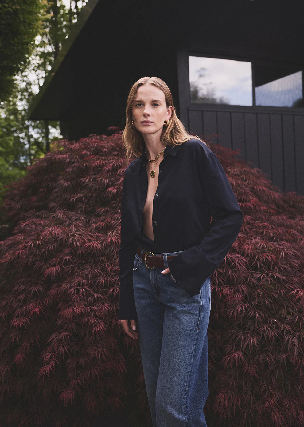 Person wearing a dark jacket and blue jeans standing in front of a building with greenery.
