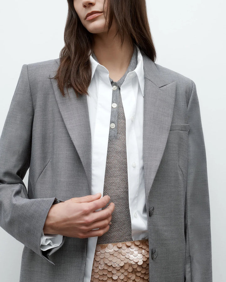 Person wearing a gray blazer over a white shirt and patterned skirt on a plain background