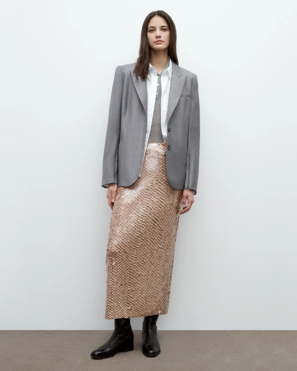 Person wearing a gray blazer, white shirt, and beige patterned skirt against a plain background