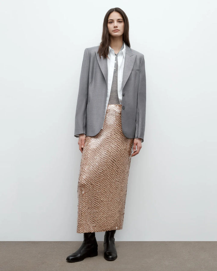 Person wearing a gray blazer, white shirt, and beige patterned skirt against a plain background