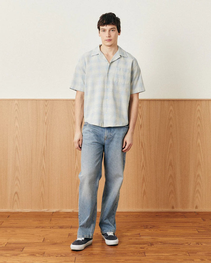 Person wearing a light-colored patterned shirt and blue jeans standing against a wooden paneled wall.