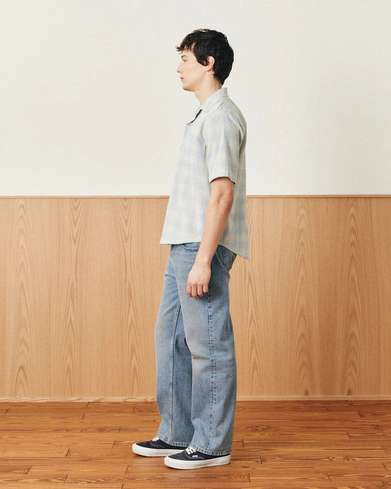 Person wearing a light-colored shirt and blue jeans standing against a wooden paneled wall.