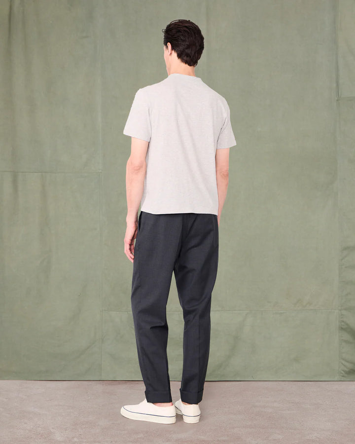 Person wearing a light gray t-shirt and dark pants standing against a green curtain.