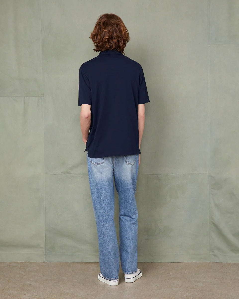 Person wearing a navy polo shirt and blue jeans standing against a plain wall.
