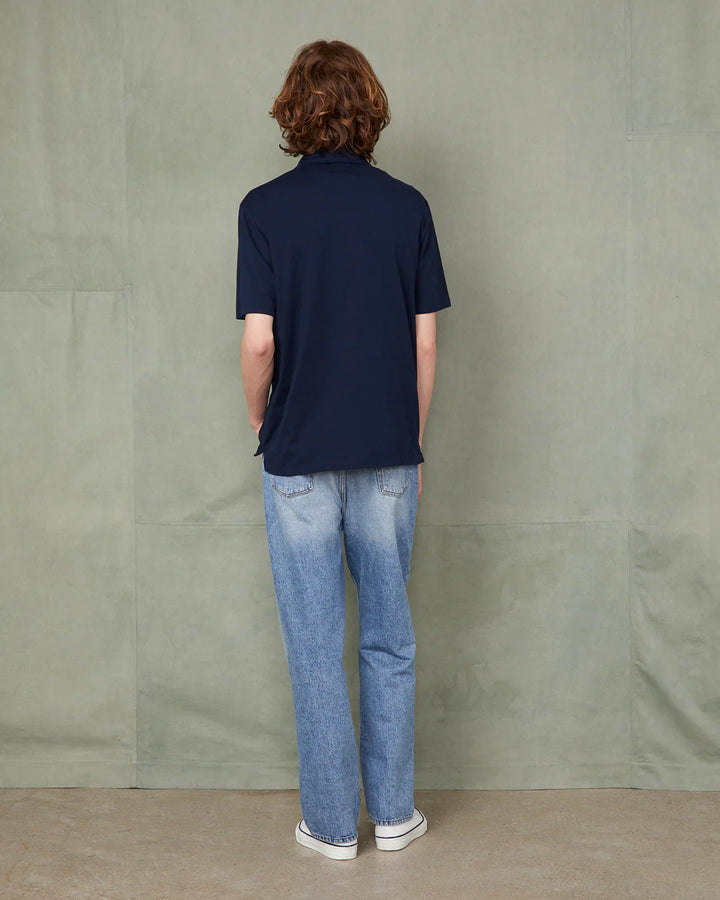 Person wearing a navy polo shirt and blue jeans standing against a plain wall.