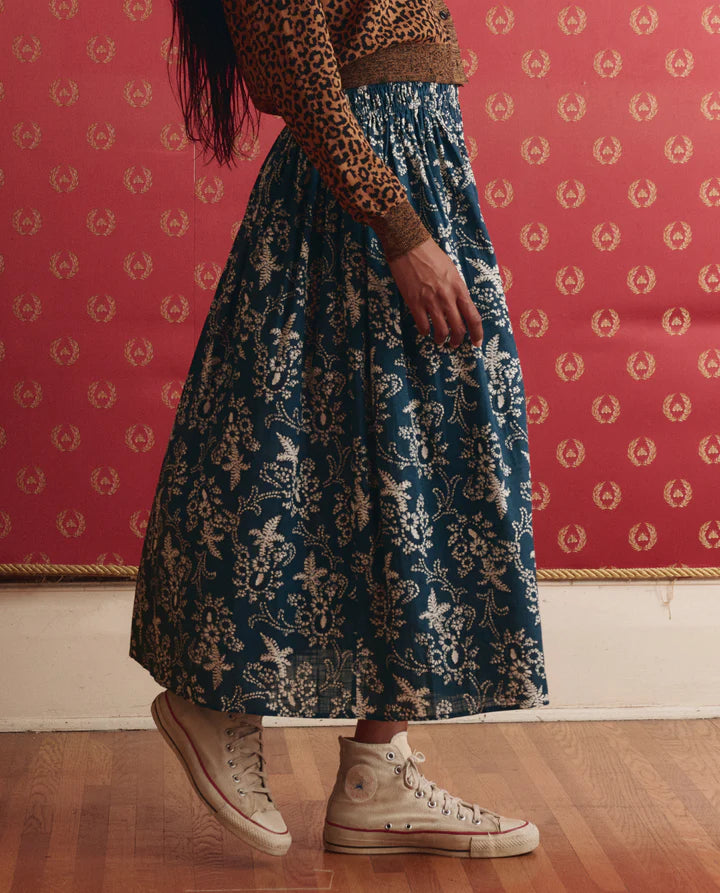 Person wearing a patterned skirt and brown sweater against a pink wall with a gold design.