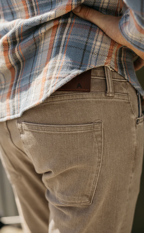 Person wearing a plaid shirt over a brown jacket with a visible brand logo, standing against a neutral background.