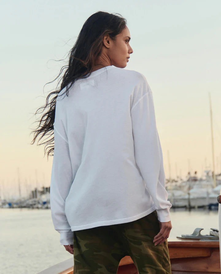 Person wearing a white long-sleeve shirt and green pants standing by a waterfront at sunset.