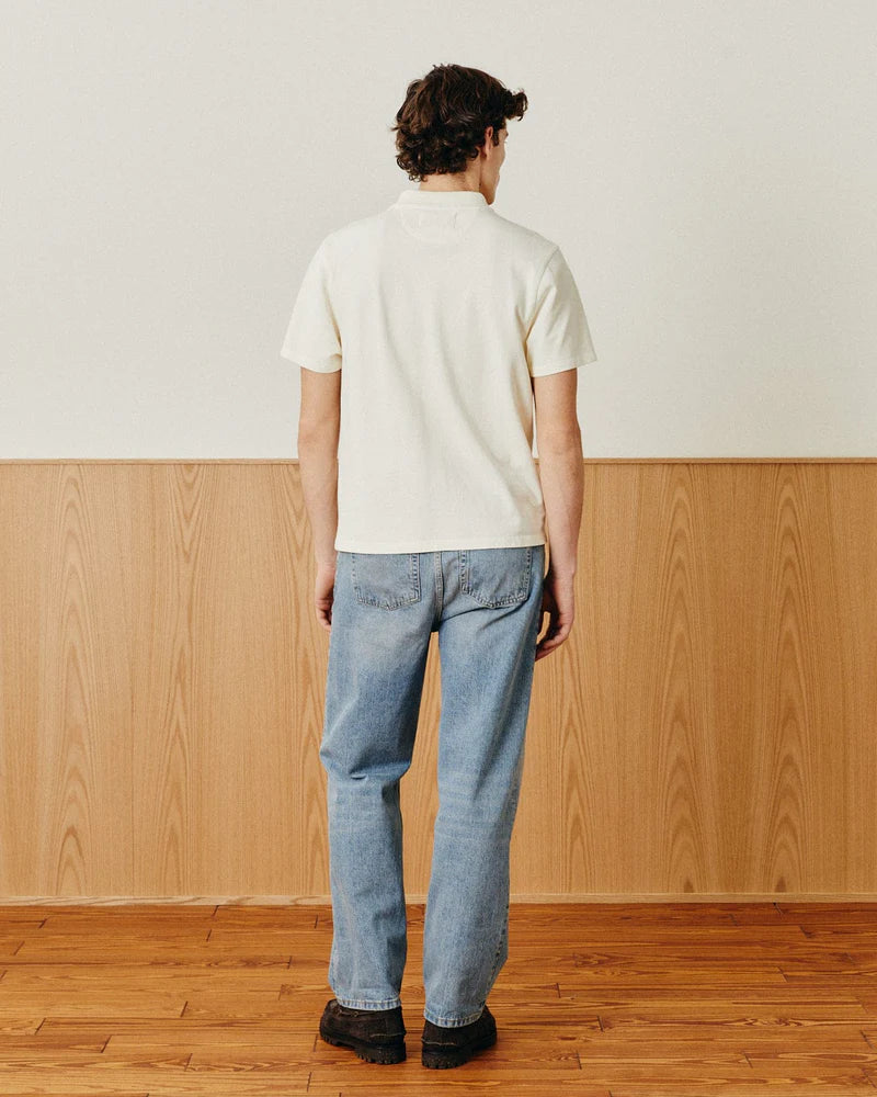 Person wearing a white polo shirt and blue jeans standing in a room with wooden flooring and paneling.