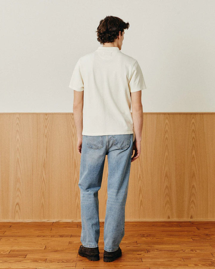 Person wearing a white polo shirt and blue jeans standing in a room with wooden flooring and paneling.