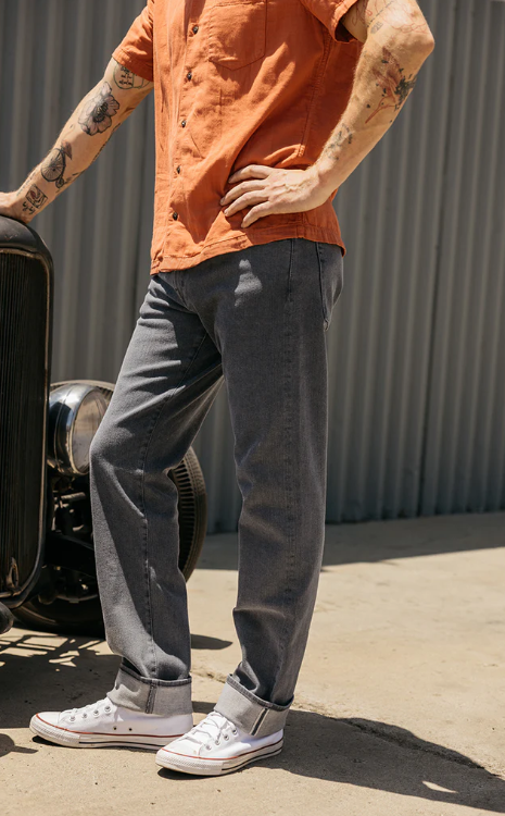 Person wearing an orange shirt, gray pants, and white sneakers standing next to a vintage car.