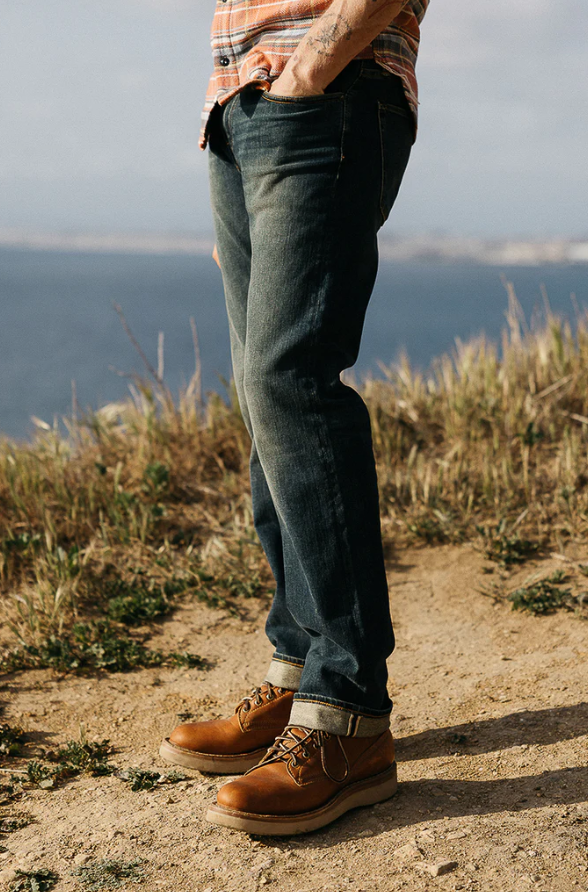 Person wearing blue jeans and brown boots standing on a dirt path with a scenic background.