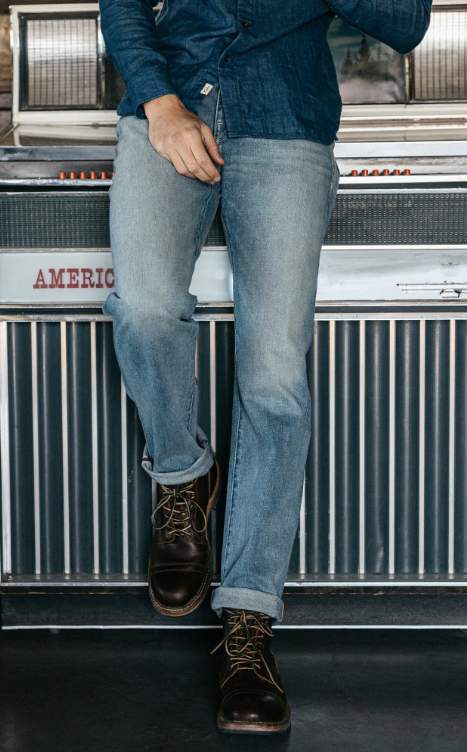 Person wearing blue jeans and dark boots standing in front of a vintage machine with 'American' branding.