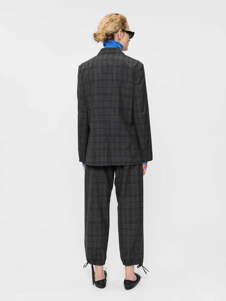A model viewed from the back wearing a dark gray and black plaid blazer and matching cropped trousers, styled with a bright blue shirt, black sunglasses, and black pointed shoes against a plain white studio background.