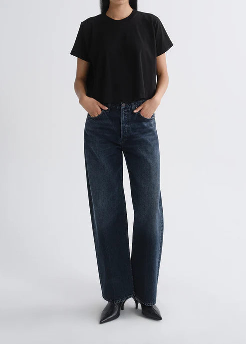 A model wearing a black short-sleeve t-shirt and dark blue wide-leg jeans, standing against a light gray studio background.