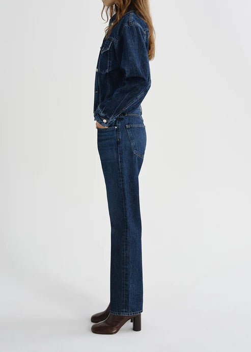 A side-profile view of a model wearing a dark wash blue denim jacket and matching straight-leg jeans, paired with dark brown heeled boots, against a plain white studio background.