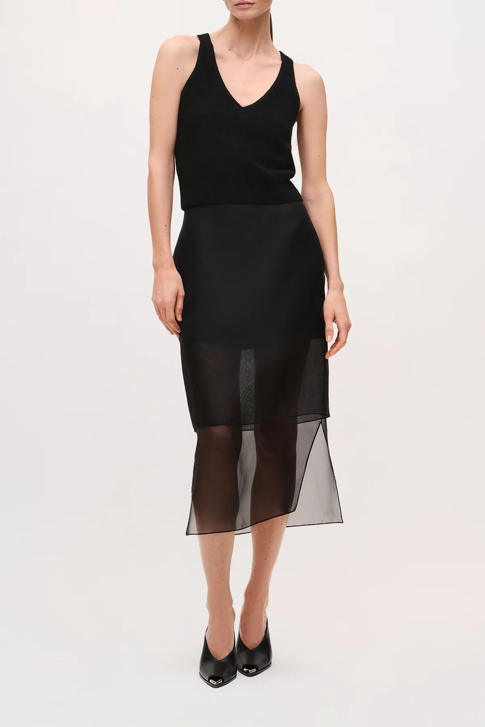 A model wearing a sleeveless black dress with a structured bodice and a sheer black midi skirt overlay, paired with black pointed-toe heels, against a plain light background.